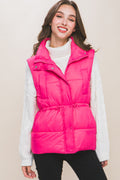 Woman wearing a bright pink puffer vest over a white knit sweater, black pants, studio background